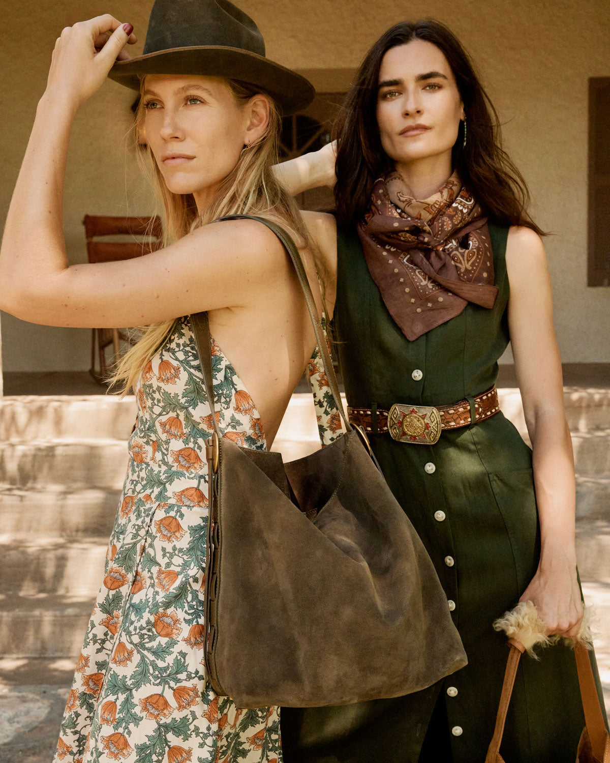 Two women standing together, one in a floral dress and wide-brimmed hat, the other in a green outfit with a scarf, against a rustic background.