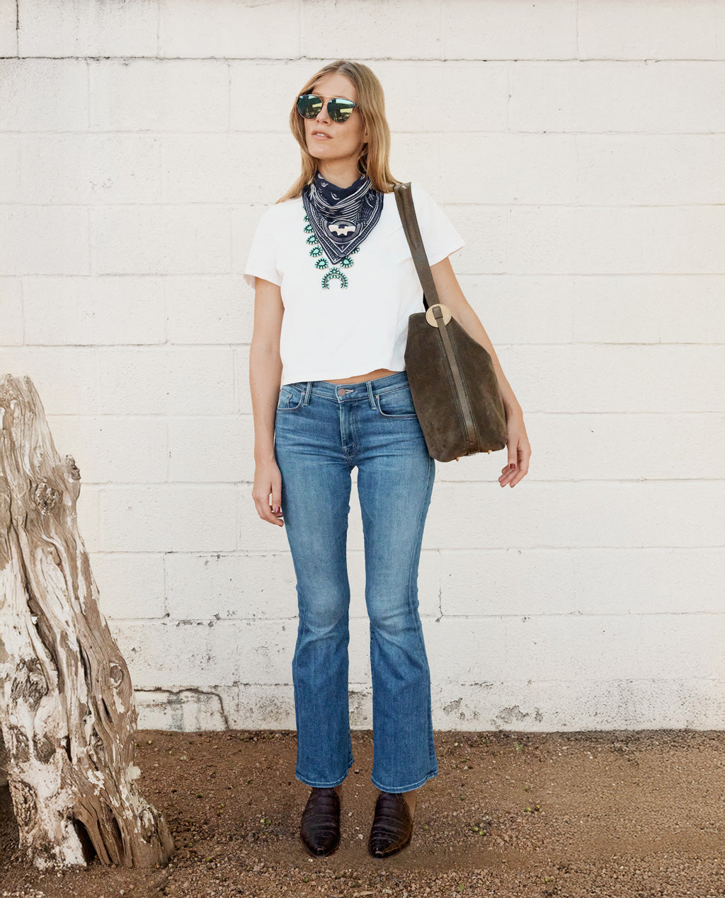 Woman wearing a white t-shirt, blue jeans, and sunglasses, standing against a white wall.