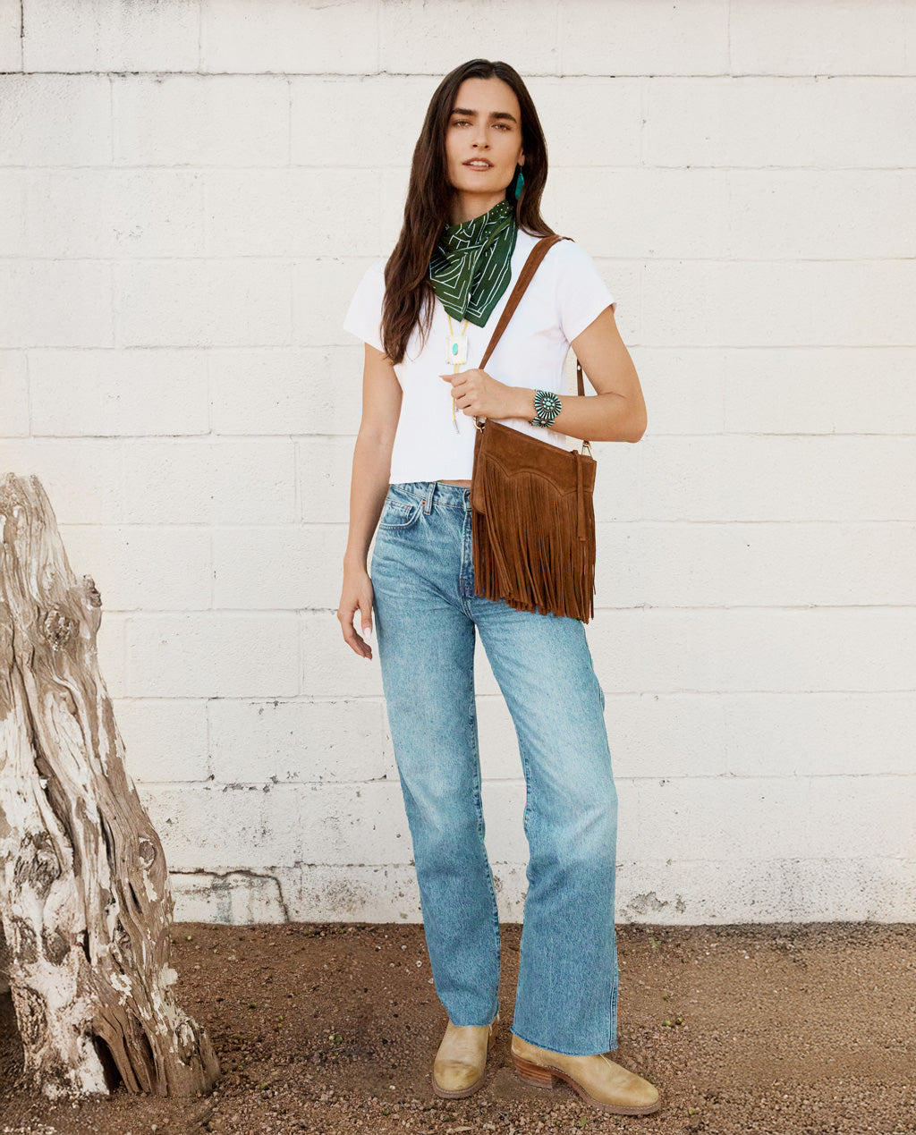 Woman wearing a white t-shirt, blue jeans, and a brown fringed bag against a white wall.