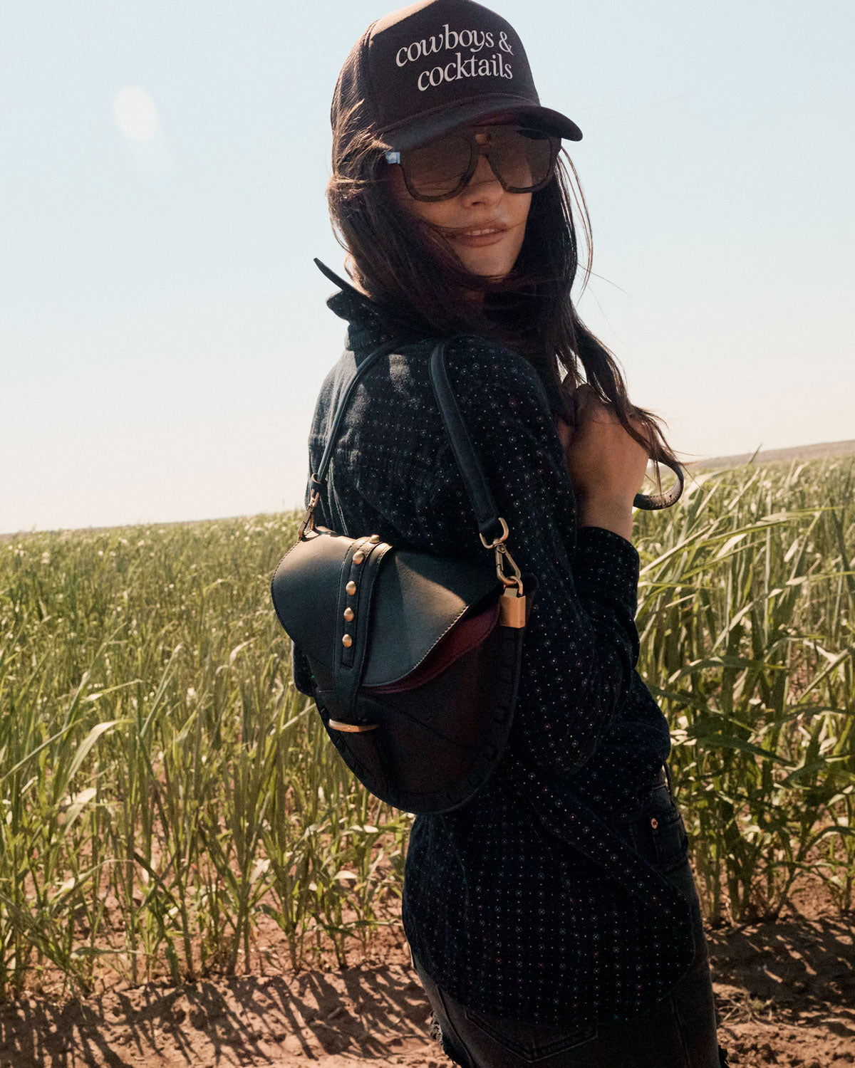 Person wearing a black cap with 'cowboys & cocktails' text, sunglasses, and carrying a black bag in a field.