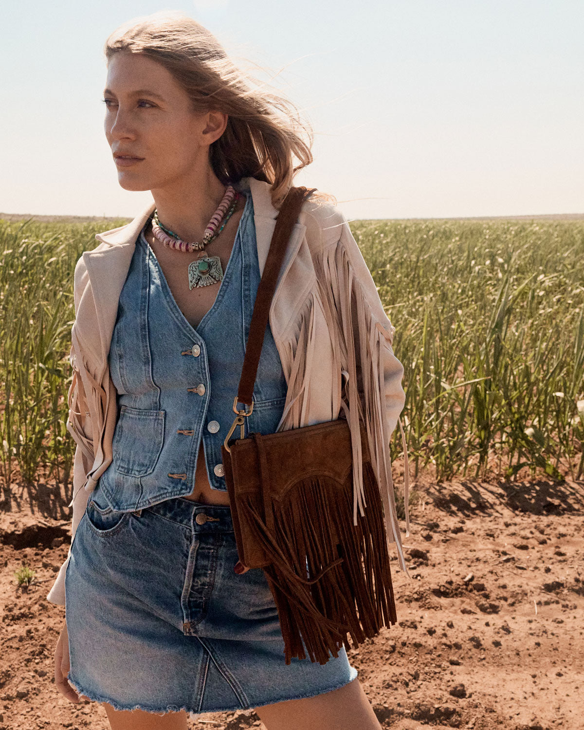 Woman in a field wearing a denim outfit with a beige jacket and brown fringed bag.