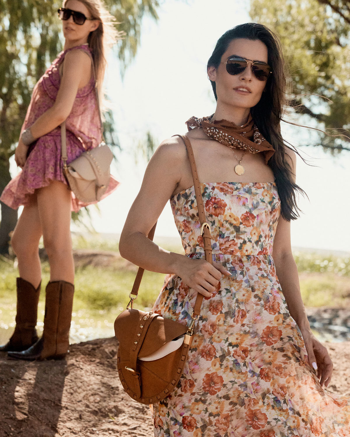 Two women in floral dresses with sunglasses and brown boots standing outdoors.