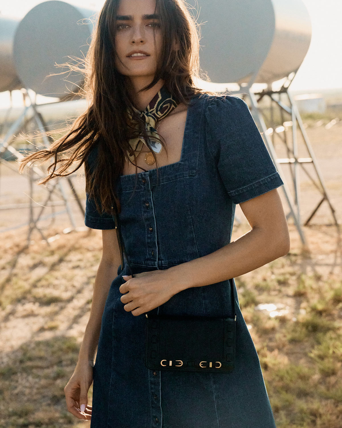 Woman in a denim outfit with a scarf, standing in an open field with large satellite dishes in the background.