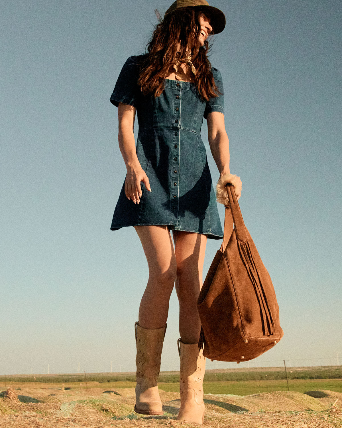 Woman in a denim dress and boots holding a brown leather bag against a clear sky.