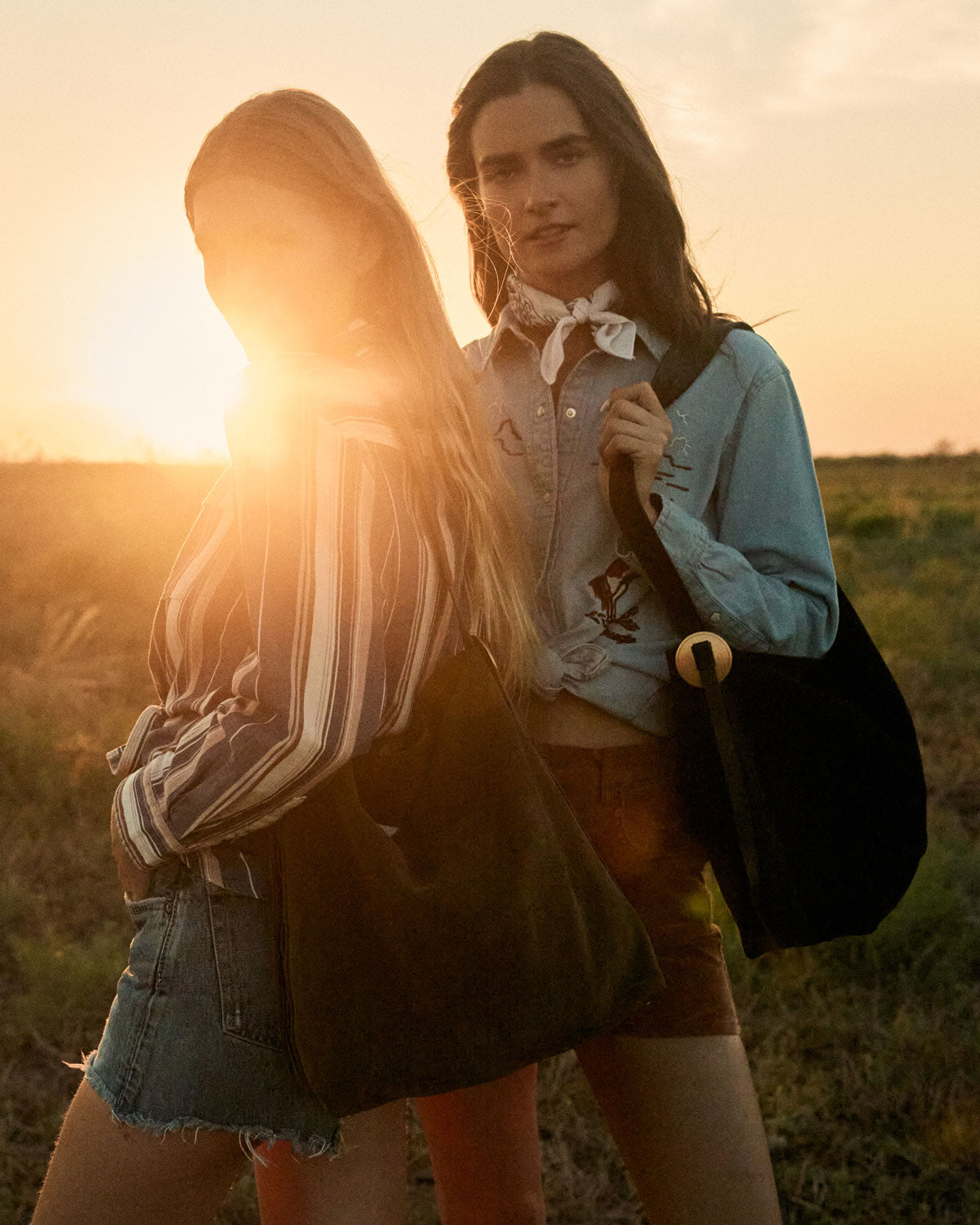 Two women standing in a field with the sun setting behind them, one holding a black bag.