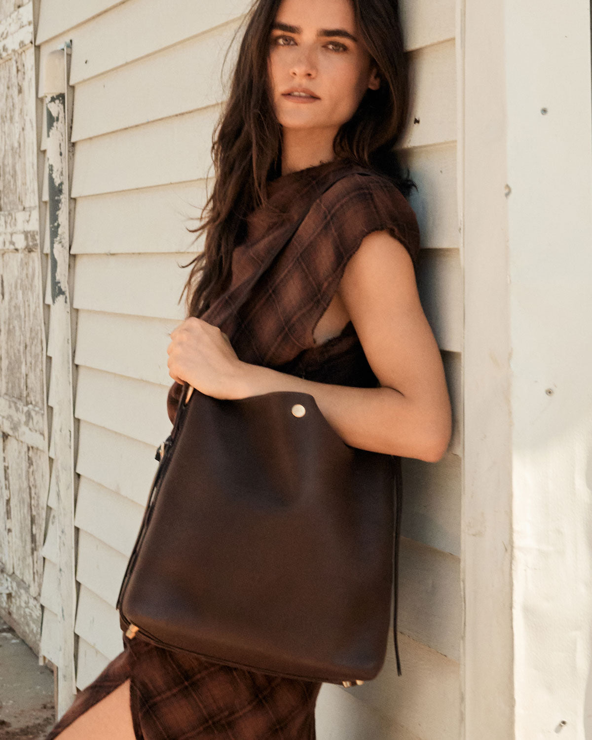 Woman holding a brown leather handbag against a wooden wall.