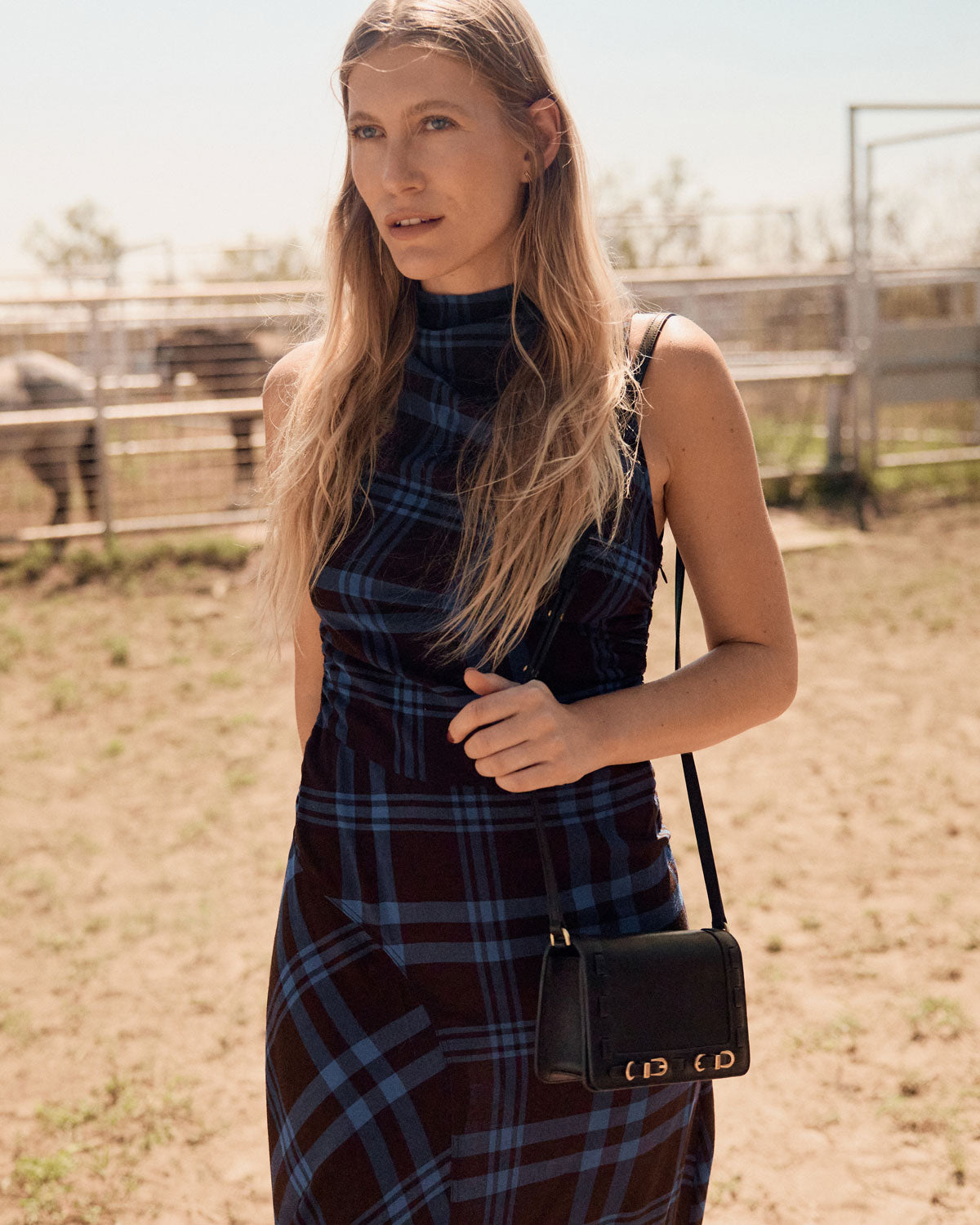 Woman in a plaid dress holding a black handbag in an outdoor setting with horses in the background.