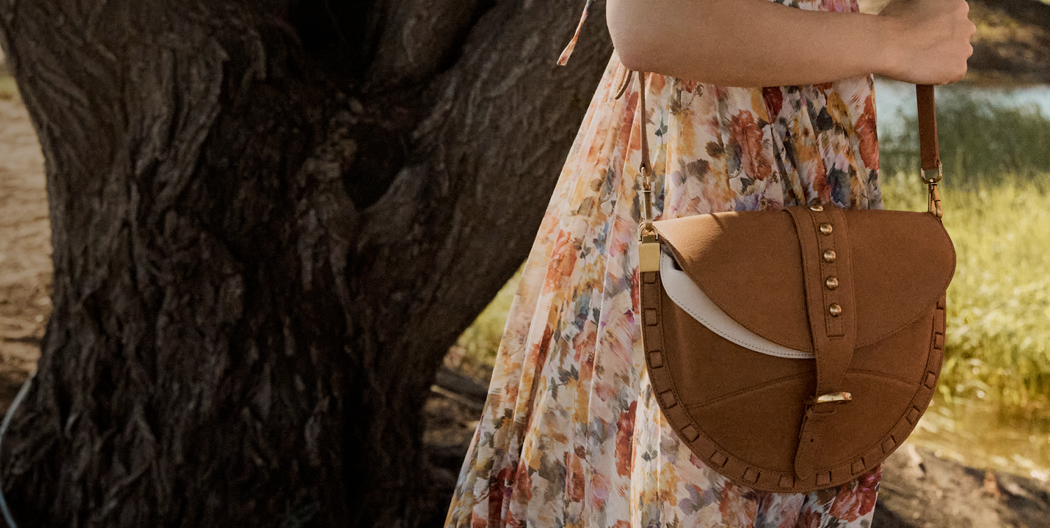 Person wearing a floral dress with a brown leather bag standing next to a tree
