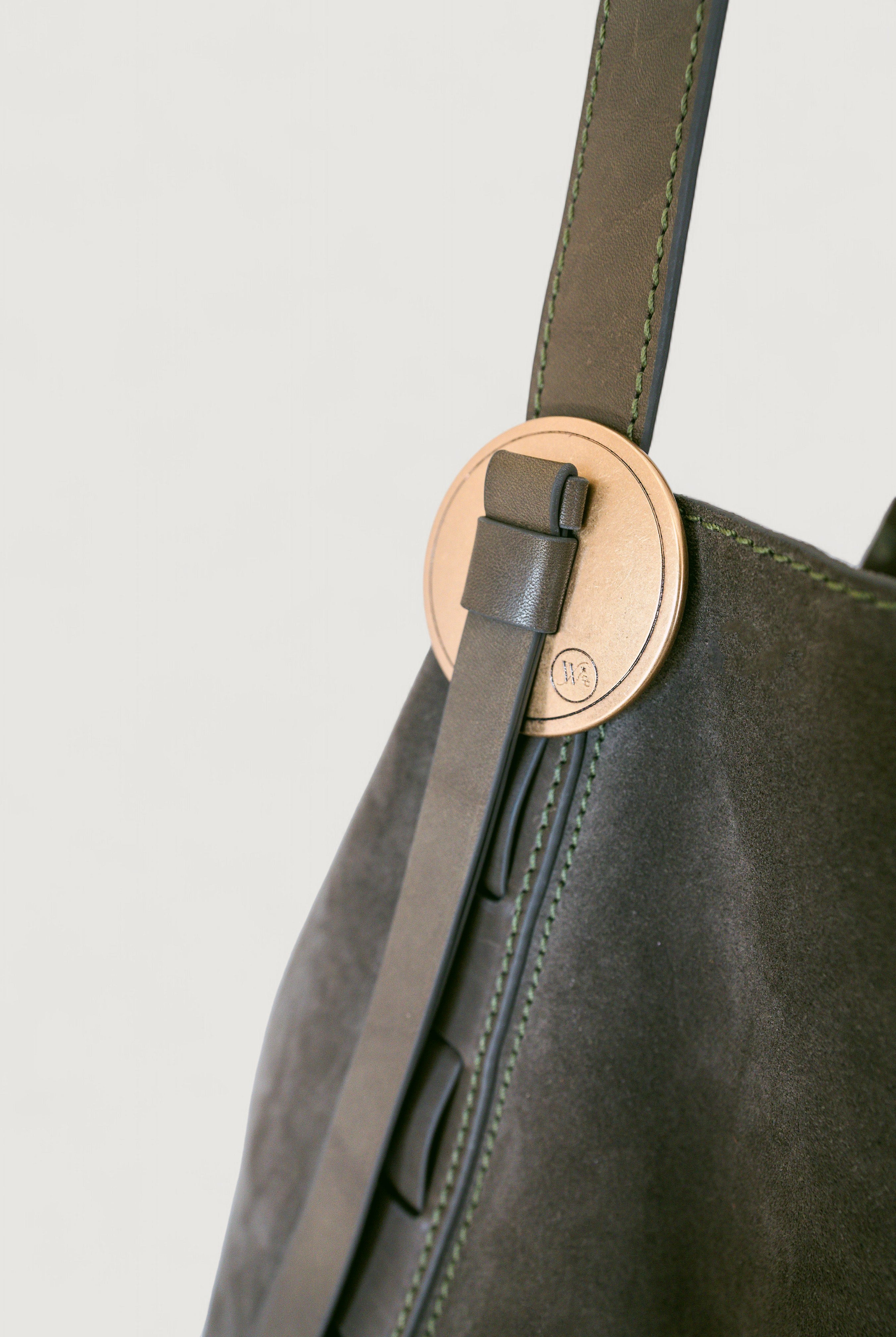 Close-up of an olive color leather bag with a strap and a brass concho on a light gray background