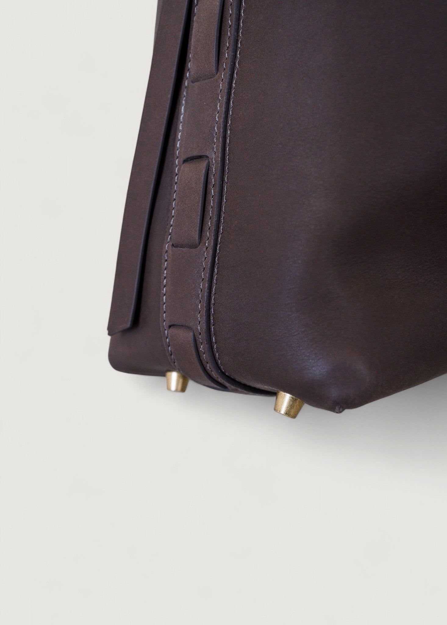Close-up of a dark brown leather bag with brass feet on a light gray background