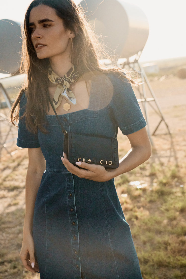 Woman in a denim dress holding a black handbag outdoors with a blurred background