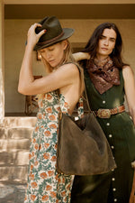 Two women in vintage-style clothing standing outdoors, one holding a brown leather bag.