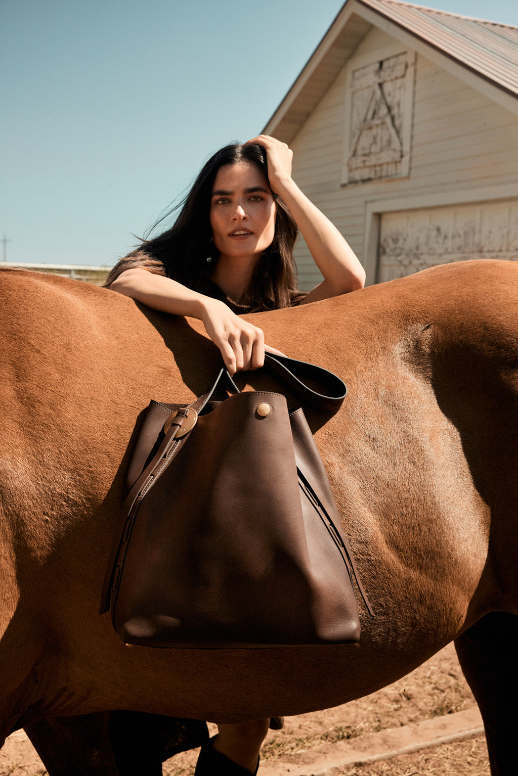 Woman holding a brown leather bag leaning over a horse in front of a barn.