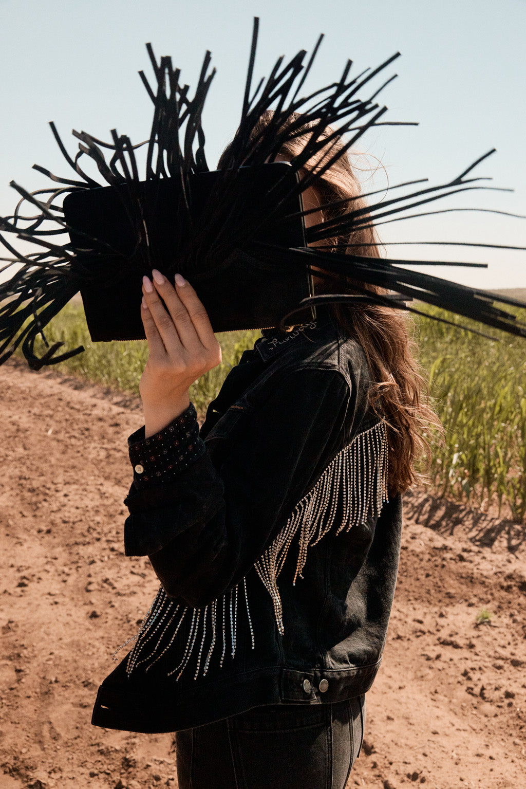 Woman holding a black clutch with long black fringe in front of their face outdoors.