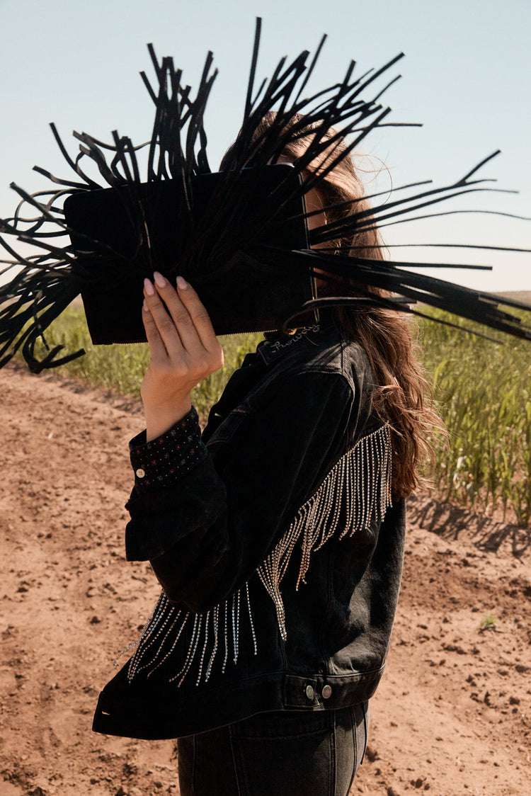 Woman holding a black clutch with long black fringe in front of their face outdoors.