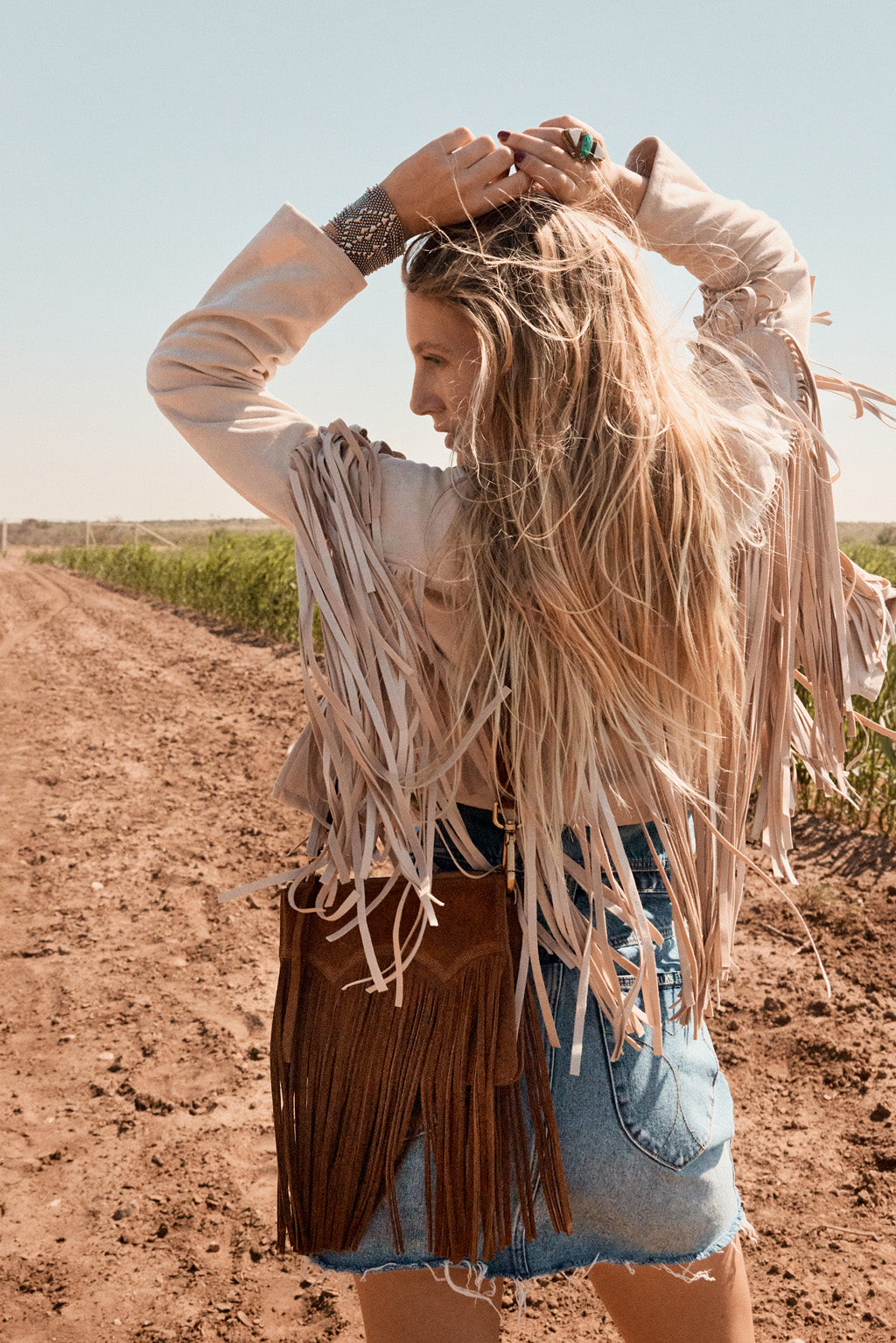 Woman with long blonde hair wearing a fringed jacket and denim shorts on a dirt road.