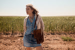 Woman standing in a field wearing a denim outfit and carrying a brown leather bag.