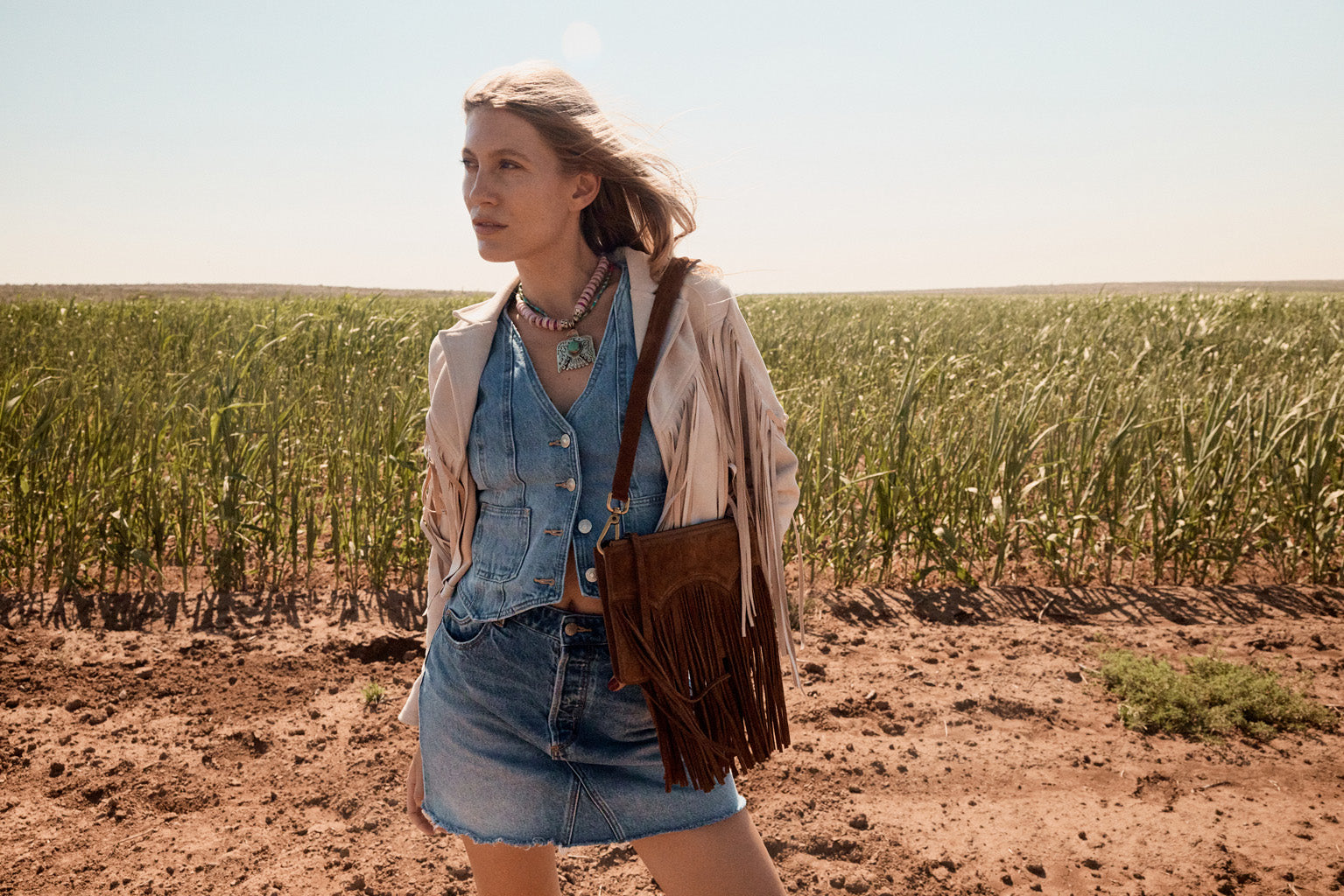 Woman standing in a field wearing a denim outfit and carrying a brown leather bag.