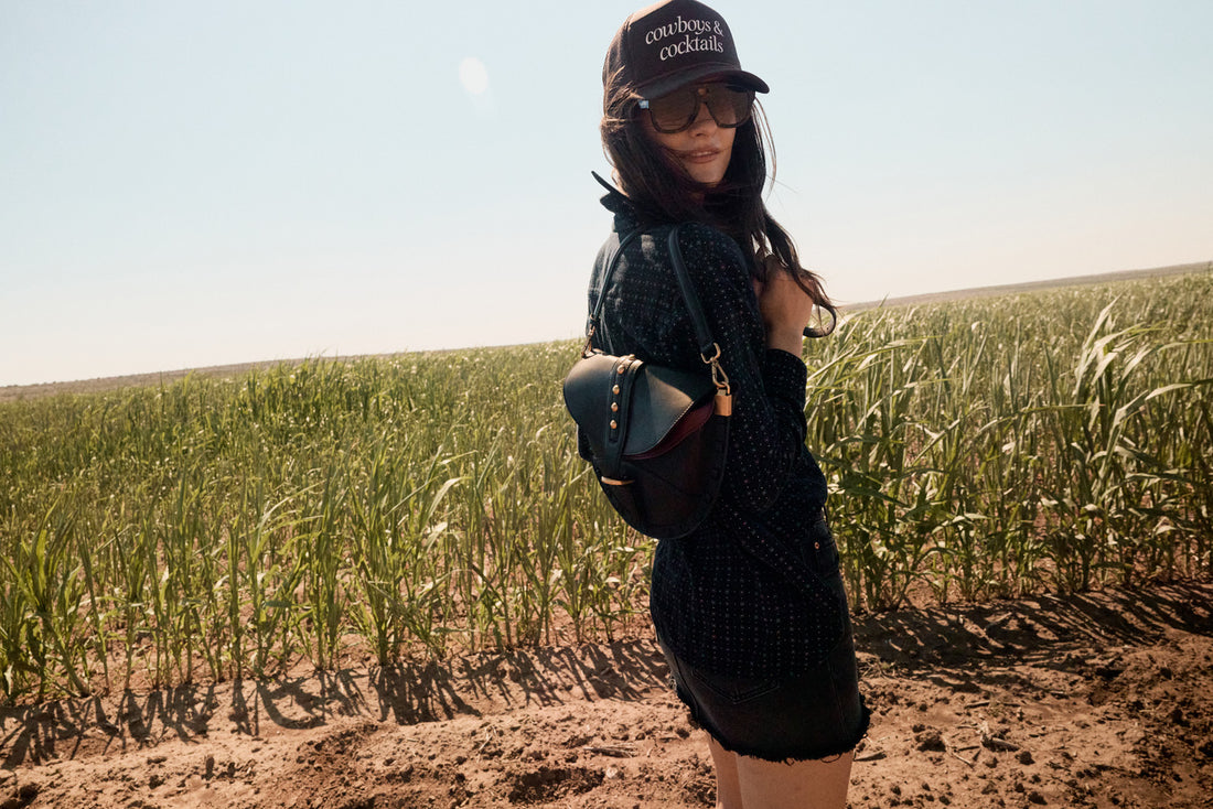 Person standing in a field wearing a black outfit and sunglasses with a clear sky.